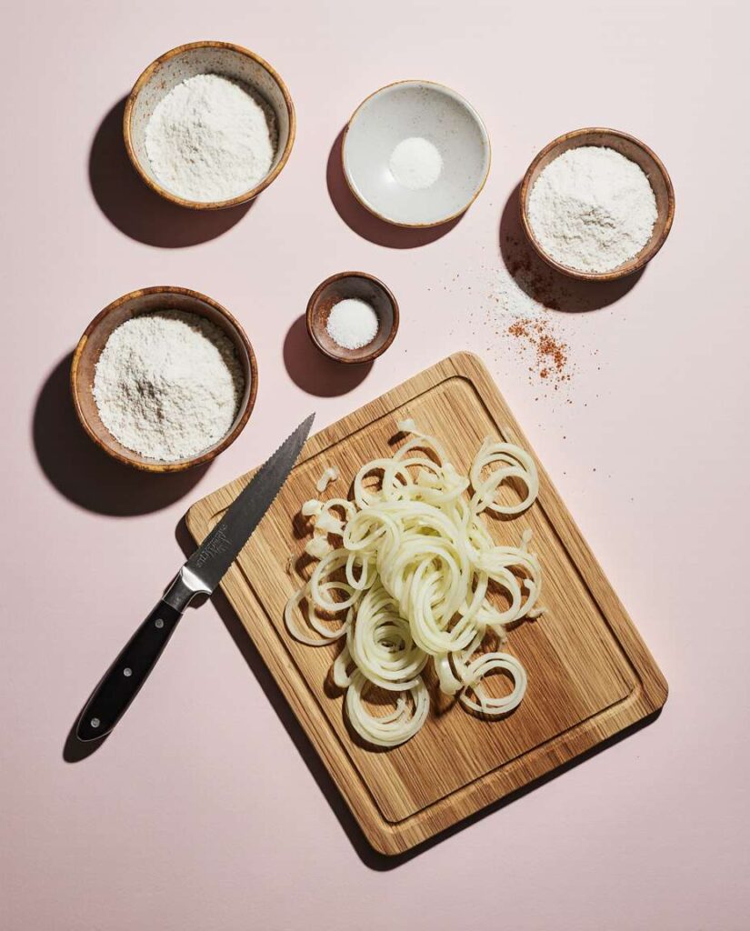 Flat lay of sliced onions, flour, salt, and cooking spray on a marble surface for air fryer crispy onions recipe.